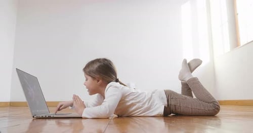 A little girl lying on the floor uses the computer and looks into the camera and smiles. Concept o