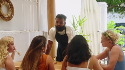 Black Waiter Talking with Female Customers at Cafe Terrace on Bali Diverse Young Women Eat Discuss