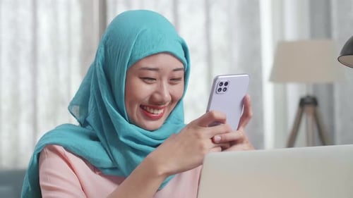 Close Up Of A Muslim Woman Smiling And Using Smartphone While Working On A Laptop At Home