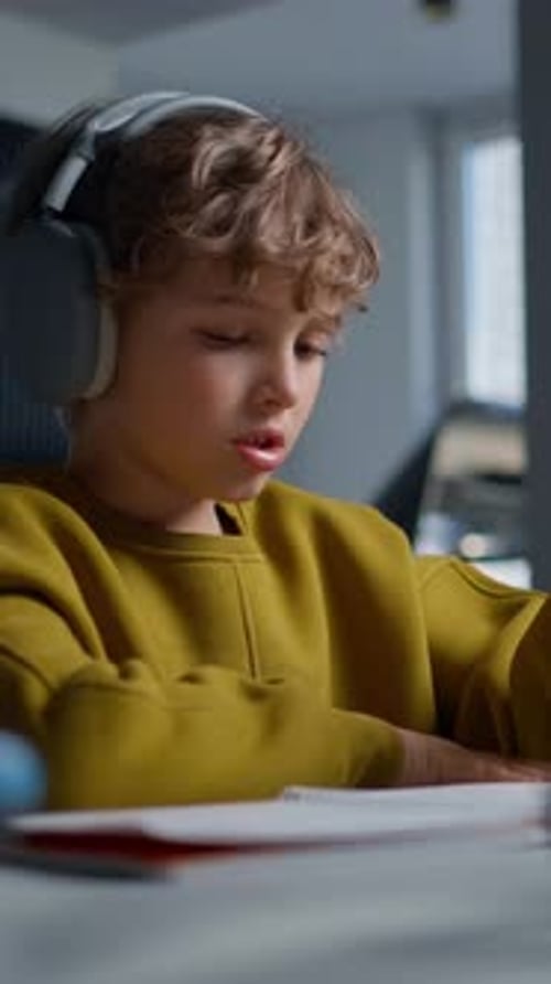 Young Boy with Curly Hair Wearing Large Headphones Intently Focused on Writing and Studying at a