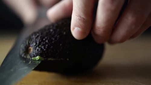 Hands of caucasian male rotating avocado on wooden chopping board while slicing it in half with shar