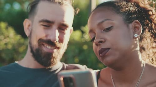 Close up, happy smiling interracial couple watching video on mobile phone while sitting on bench
