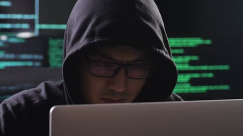 Close Up Of Asian Male Hacker In A Hood Works On A Laptop With Maps And Data On Display Screens