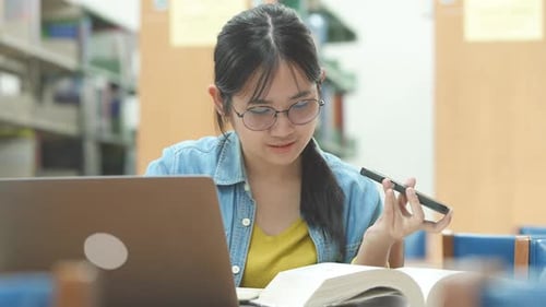 Asian student doing research in library using laptop books making a project and preparing for exam.