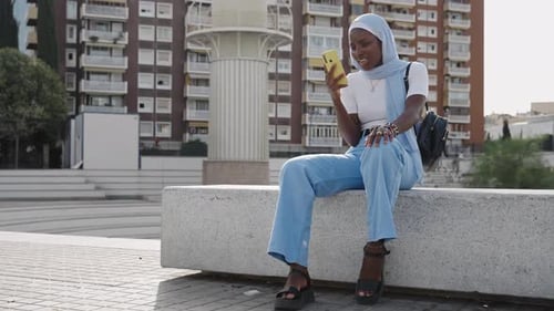 Young Muslim Woman with Blue Hijab Using Yellow Cell Phone App in the City