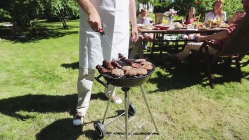 Food, People And Family Time Concept - Man Cooking Meat On Barbecue Grill At Summer Garden Party