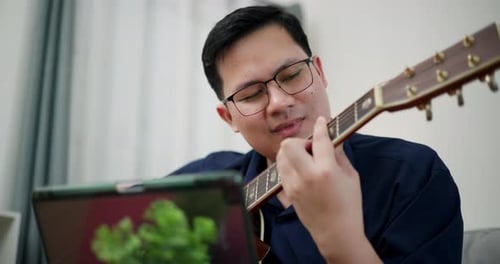 Homme asiatique détendu jouant joyeusement de la musique avec une guitare sur le canapé à la maison.