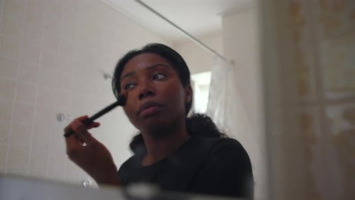 African American woman blending makeup on cheek with brush while looking at her reflection in