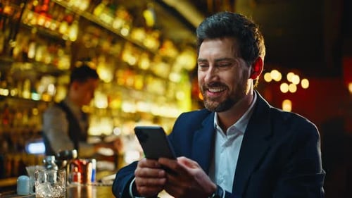 Happy Man Savouring Whiskey Looking Mobile Waiting in Glowing Lounge Closeup