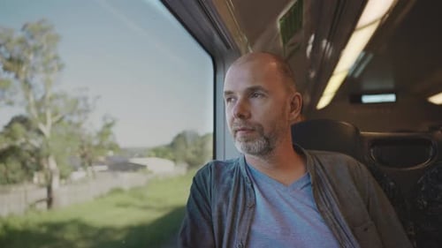 A Middleaged Bald Man in a Shirt Sits By the Train Window Gazing at Passing Rural Landscapes During
