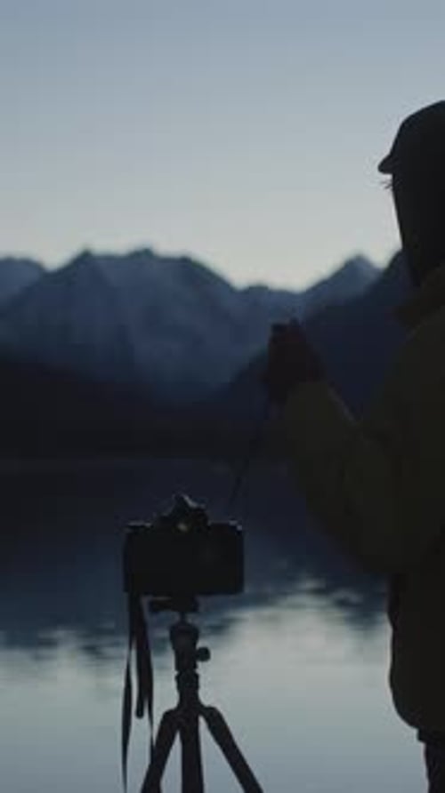 Photographer with Camera on Tripod By Mountain Lake at Dusk