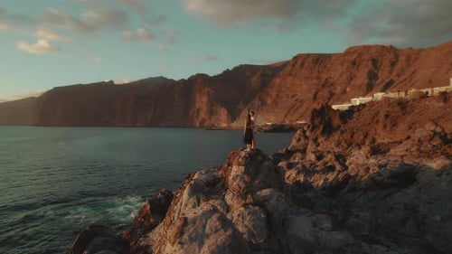 The couple stands on a rocky cliff by the ocean, the setting sun casting a warm glow over them as th