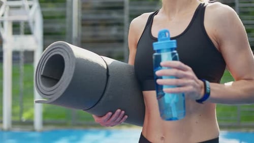 Slender Female Moving Slowly with Yoga Mat and Water Bottle Through Stadium Area