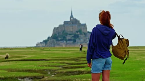 A Female with a Backpack Visits the Mont Saint Michel Castle While Walking Through Green Meadows