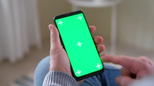 Young Man Sitting at Home Holding Smartphone Green Mockup Screen in Hand Male Person Using Chroma