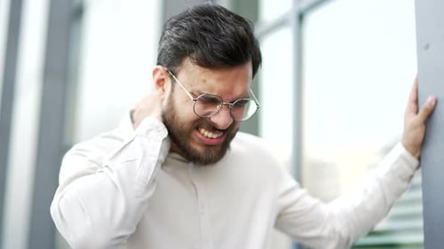 Tired businessman suffering from neck pain while standing on the street near a modern office buildin