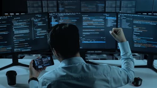 Happy Asian Man Looking At Code On Mobile Phone While Write Code With Multiple Screens In office