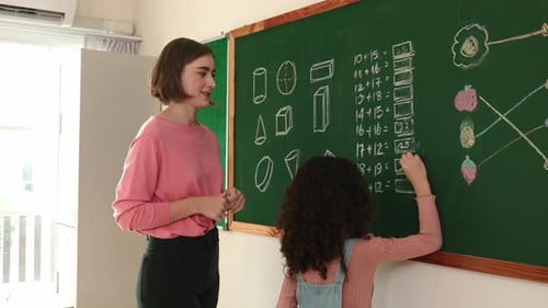 Skilled teacher looking at african girl writing answer on blackboard. Pedagogy.