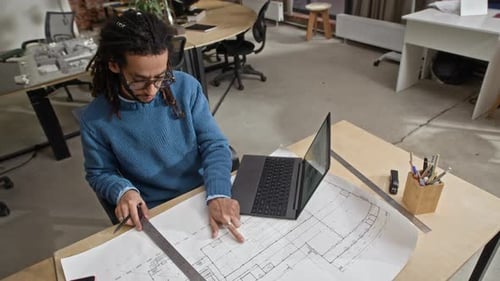 Architect Working on Blueprints with Laptop at Desk