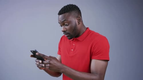Goodlooking Young Bearded AfroAmerican Man Actively Uses His Smartphone for Texting Messages on Grey