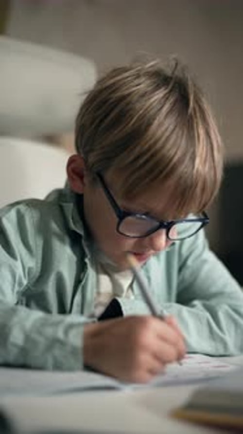 A Child Wearing Glasses Sits at a Table and Works on Exercises in a Copybook