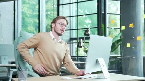 Overworked tired young business man employee with glasses with severe back pain hurt after hard work
