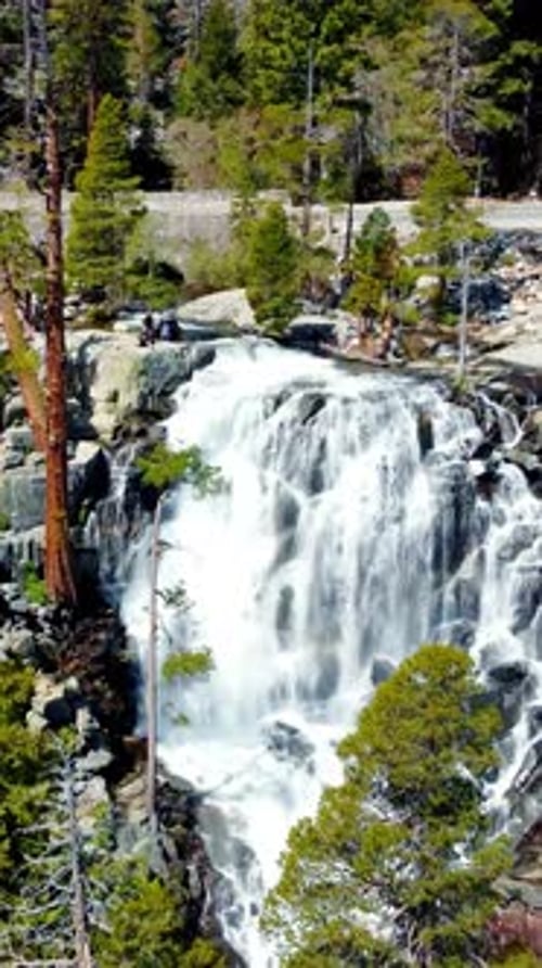 Drone Approaches a Waterfall Pouring From Stone Cliffs Into the Valley Beside a Forest Road Green