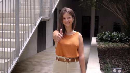 Young White Woman in Orange with Thumb Up Standing Outside University Patio Looking and Smiling