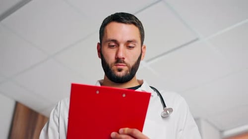 A doctor in a lab coat with a stethoscope in a patient’s room fills out documents in folder