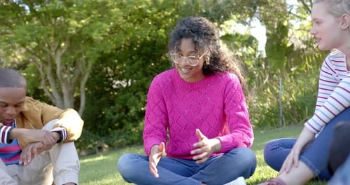 Happy diverse group of teenage friends sitting on grass and talking in sunny park, slow motion