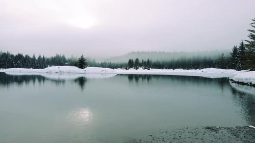 Low Drone Flight Above Alpine Lake In Snowy Mountain Forest With Hazy Fog Clouds
