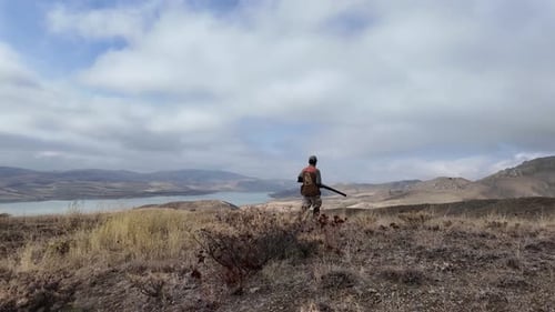 Hunter Walking Through River Valley in Mountain Landscape
