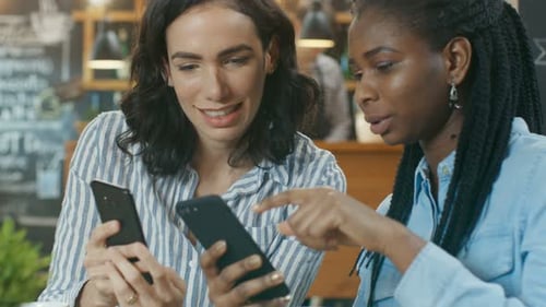 Two Gorgeous Girlfriends Have Fun while Sitting in the Cafe, They Show Each Other Something on Thei