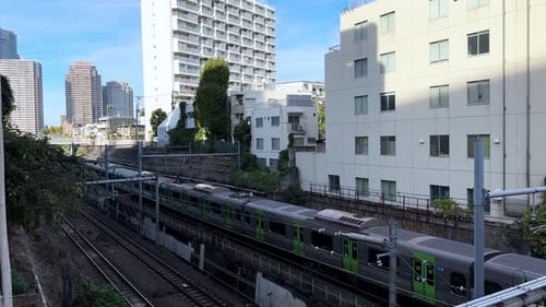 A green commuter train moves through a modern urban area with tall buildings in Tokyo, Japan