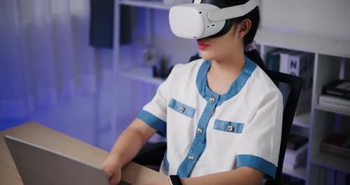 Asian woman using virtual reality headset while typing work on laptop in modern workspace.