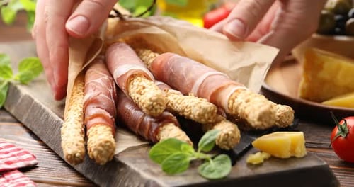 Femme prenant un gressin de grissini avec du prosciutto sur une planche à une table en bois, gros plan