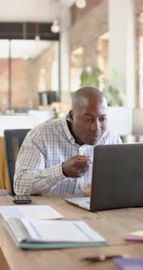 Vertical video of african american casual businessman using laptop and having lunch, slow motion