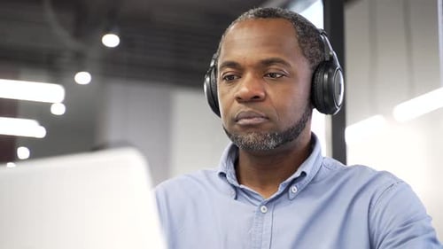 African american IT specialist in wireless headphones works on a laptop sitting at a workplace