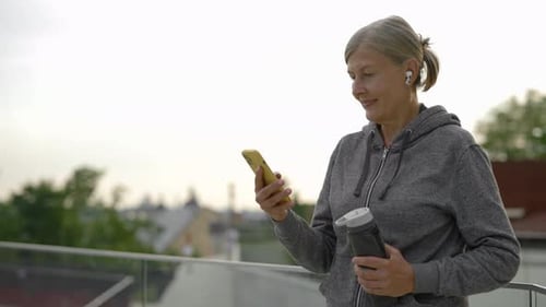 Senior Woman Using Smartphone Wearing Earphones and Holding the Sport Bottle Standing in the Park