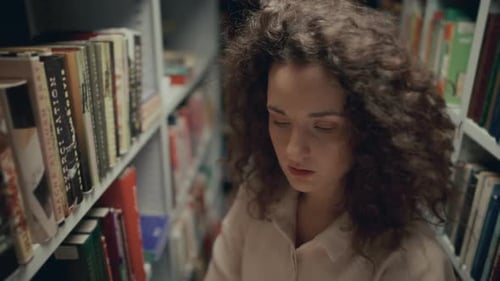 Young Female Student Picking Up Book from Shelf in Library