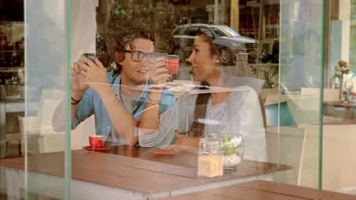 Wide Shot Through Window Of Young Couple Using Smart Phone In Cafe.