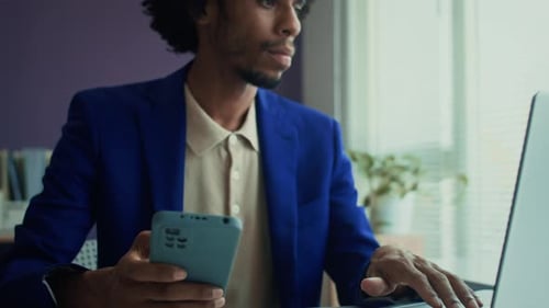 Male Entrepreneur Filling Report on Laptop Using Smartphone at Office