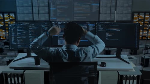 Vista trasera de un hombre asiático cansado estirándose mientras escribe código con múltiples pantallas de ordenador en la oficina