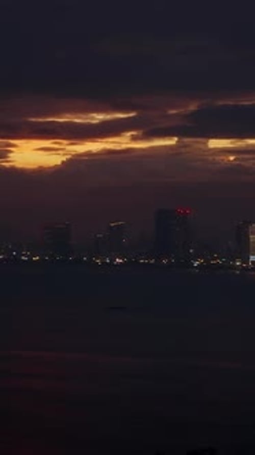 Vertical video. City Skyline Lights Reflecting on Dark Water beneath Fiery Sunset Clouds in Wide