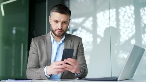 Serious businessman is using smartphone sitting at workplace in business office. Thoughtful handsome