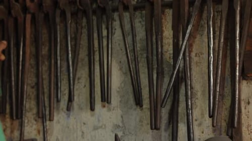 A blacksmith selects a pair of tongs from his workshop's wall, test them on a piece of metal.