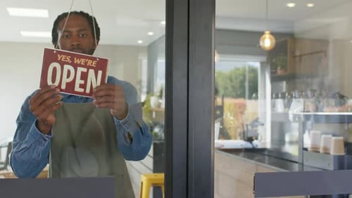 African American male barista opening coffee shop unhooking sign flipping to open peering outside