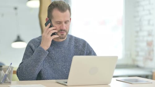Man Talks on Phone While Using Laptop at Desk