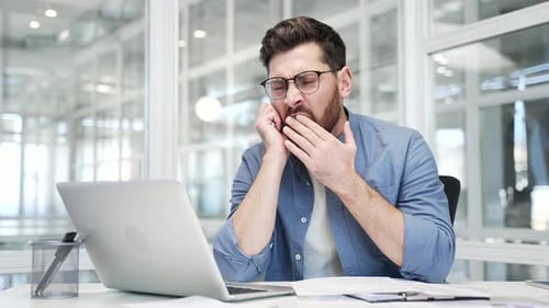 Sleepy businessman is bored sitting at workplace at desk in business office. Man yawns,