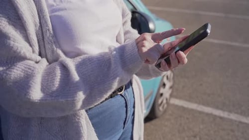Anonymous woman leaning on car using smartphone in sunny parking lot